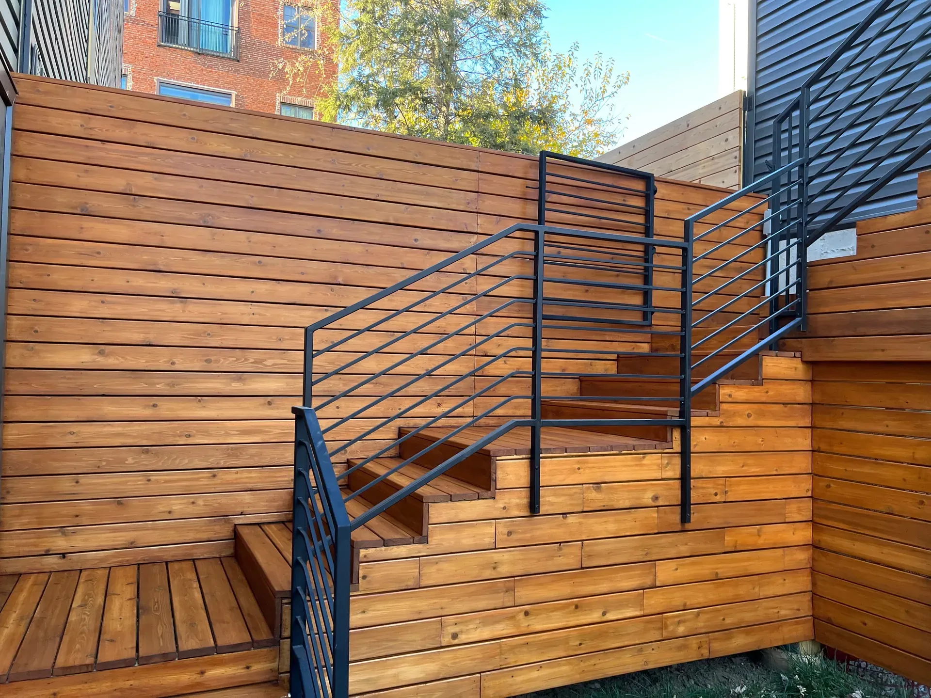 Finished cedar tongue-and-groove exterior stair and surround with black powder-coated steel railings, urban rowhouse setting, late-afternoon sun on the cedar