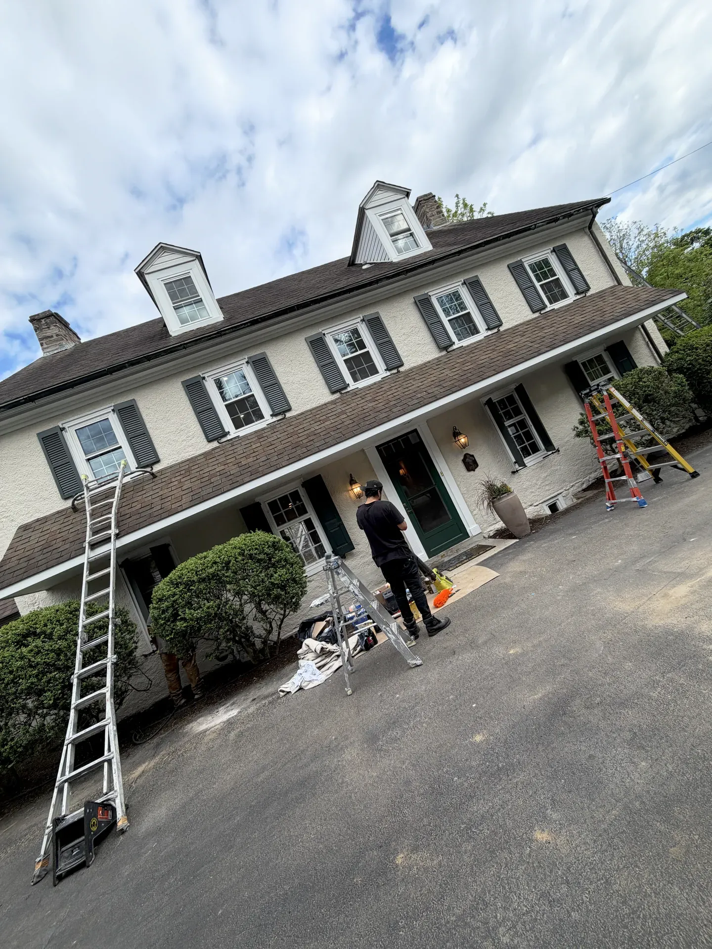 JL crew member working off a step ladder at the front entry of a large white Pennsylvania colonial mid-repaint, full-height extension ladders propped against the eaves on both ends of the house, crew supplies staged in front of the dormered facade