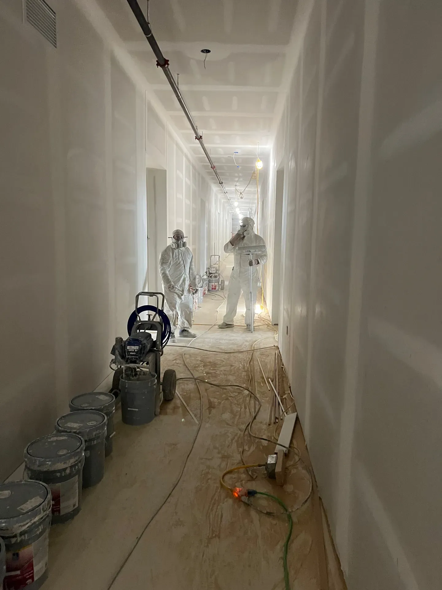 Long commercial corridor mid-drywall finish — two JL crew members in white Tyvek suits and respirators with an airless spray rig, fresh white walls under construction lights, taping compound buckets staged along the wall