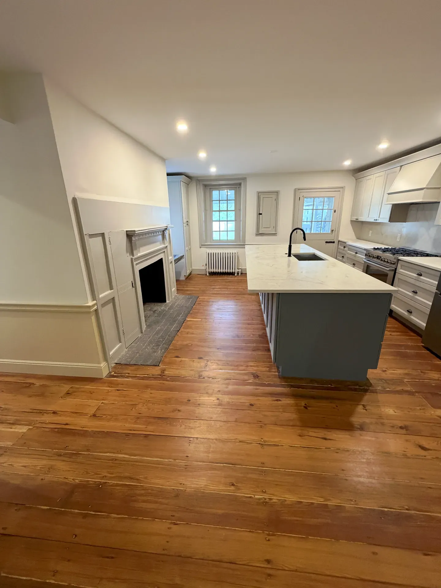 Modern kitchen with freshly refinished wide-plank pine floors, white shaker cabinets, fireplace mantel, and a large island with quartz top and bowl sink
