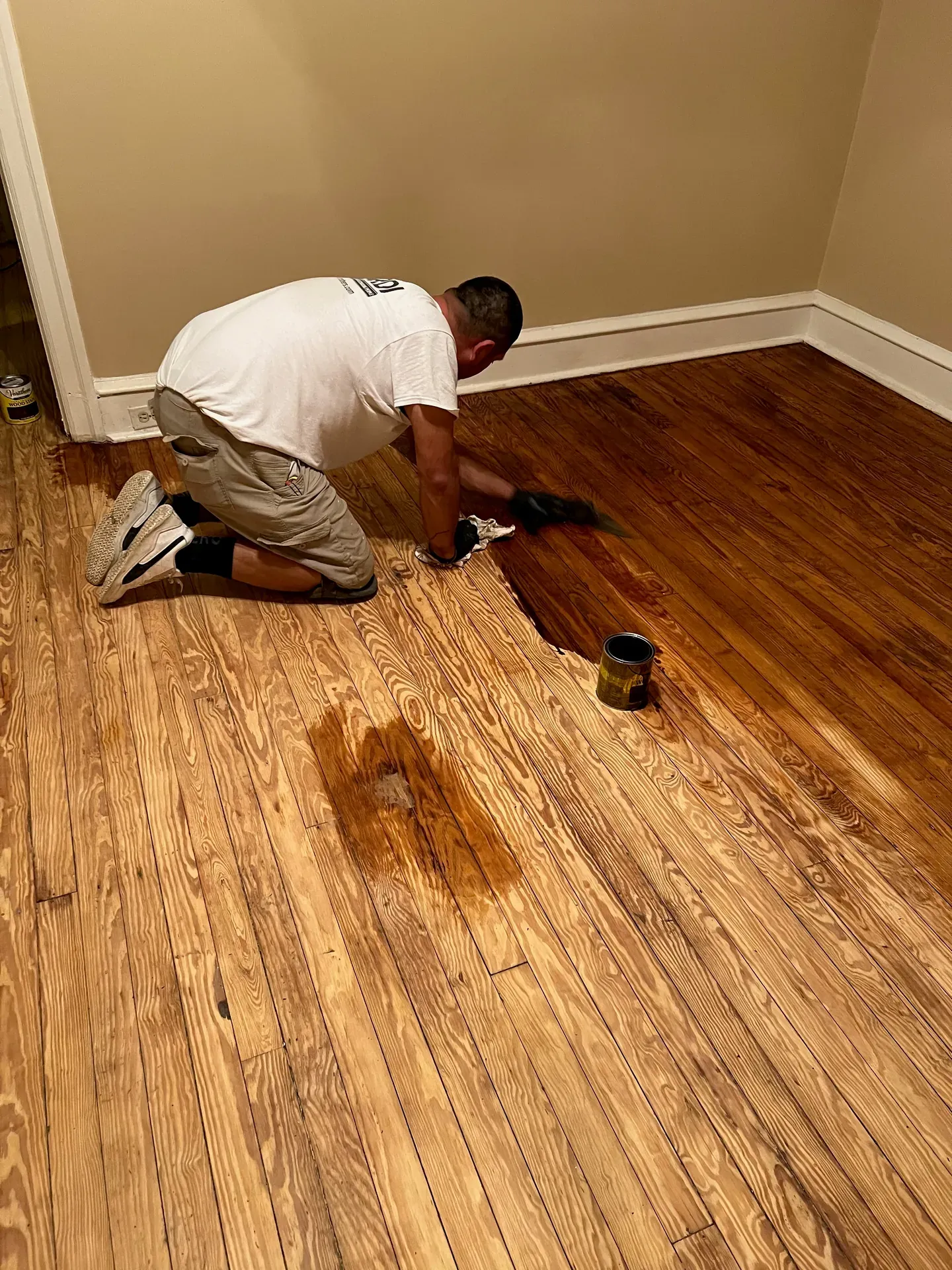 JL crew member on knees applying dark stain to a wide-plank pine floor — half the floor stained in a dark walnut tone, the other half still showing the raw sanded pine, brush and stain can on the floor next to the worker