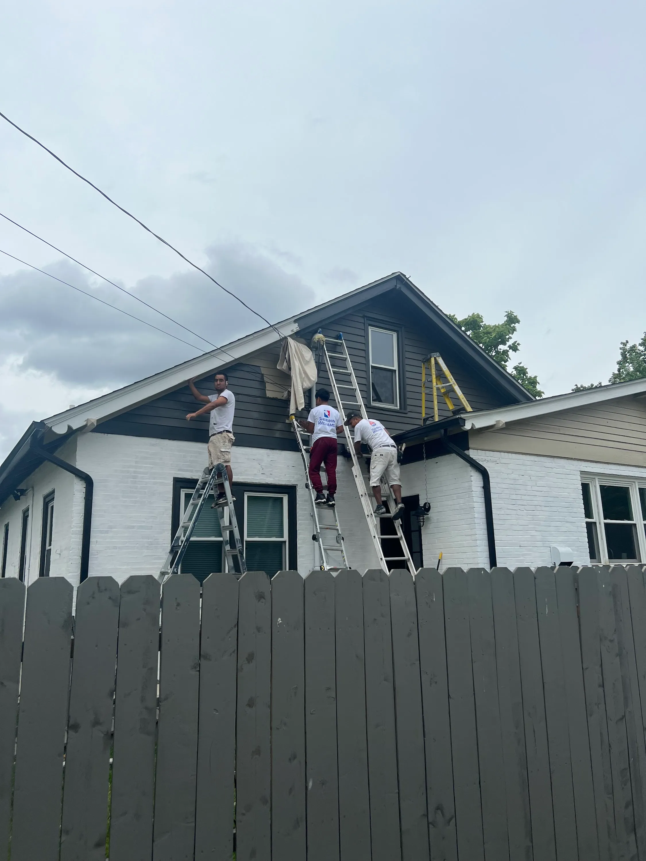 Three JL crew members in branded white tees working on a small bungalow's dormer trim, three extension ladders against the front, summer day