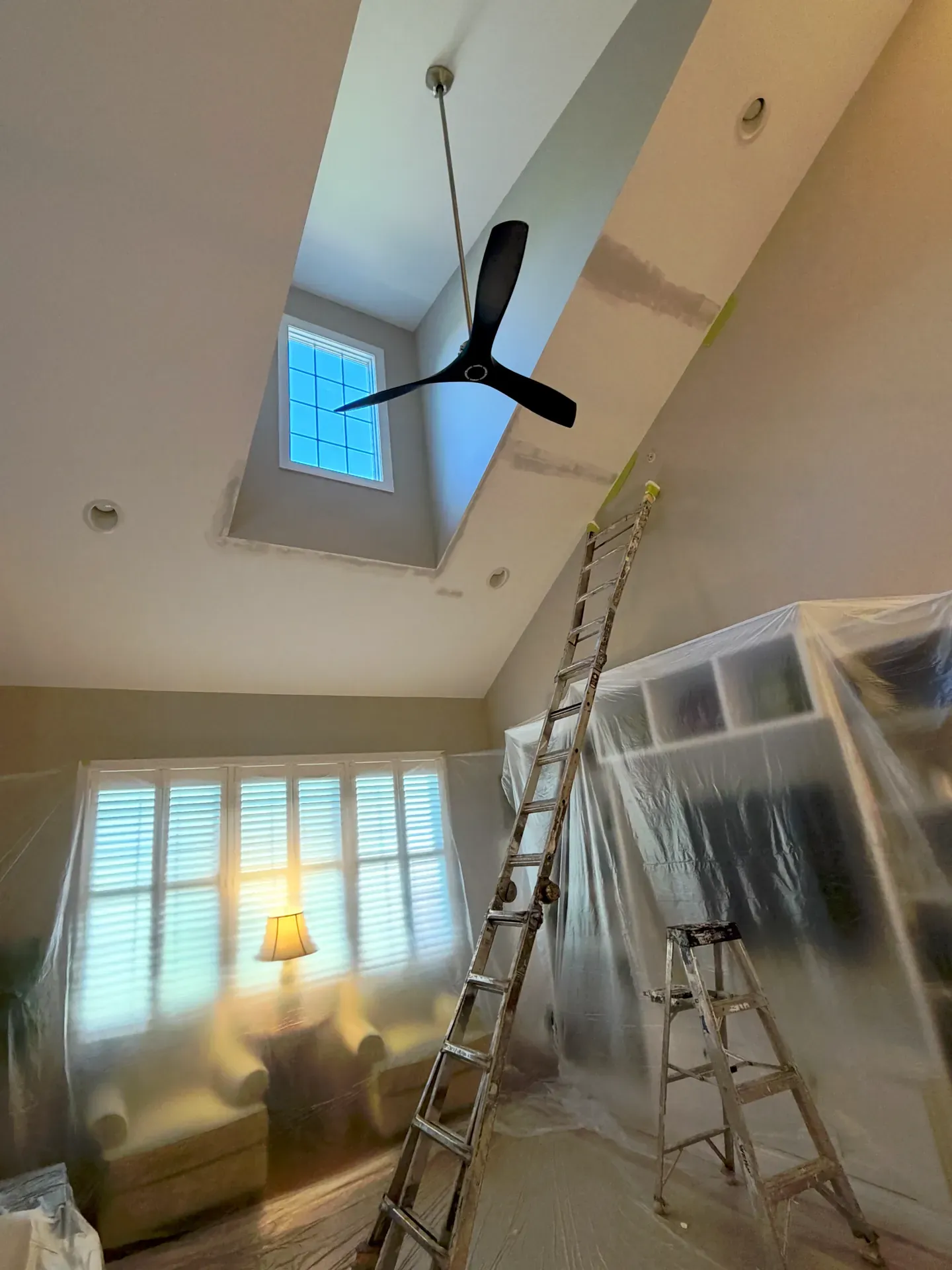 Vaulted residential ceiling mid-paint with the ceiling fan in place, ladder and step ladder set up, plastic sheeting protecting the shutters and furniture below